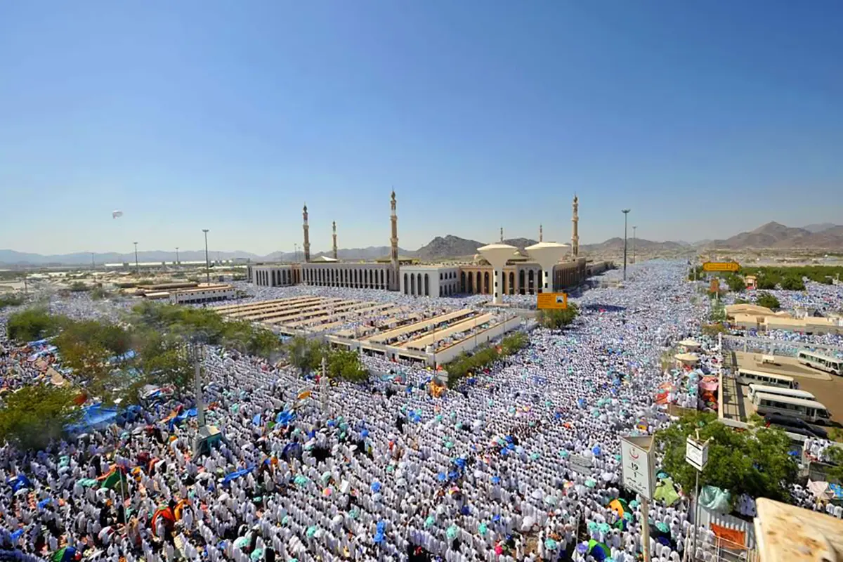 A shallow dry watercourse running along the western edge of the Arafat plain, pale gravel and low banks, boundary markers standing at intervals and the long wall of Masjid Namirah beyond.