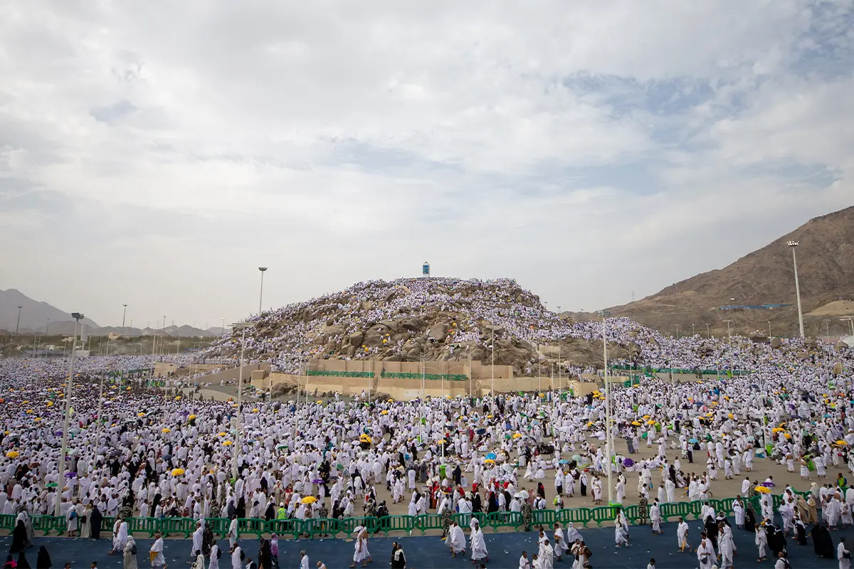 The low rocky hill of Jabal al-Rahmah rising out of the flat Arafat plain, its white stone pillar standing on the bare summit, worn steps climbing the near face and open gravel ground all around.
