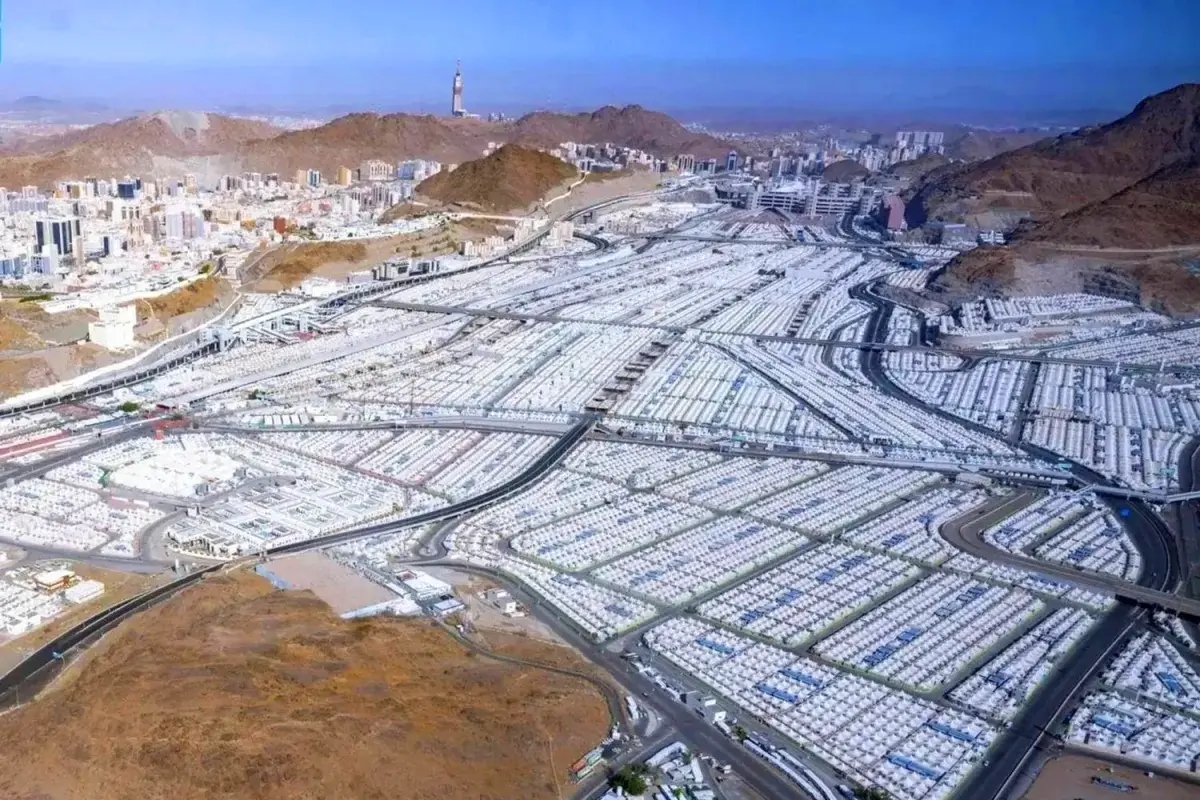 Bare rock and scree on the mountainside above the Mina valley, a shallow hollow in the stone catching the low morning light, Hajj roads and tent blocks laid out on the flat ground below.