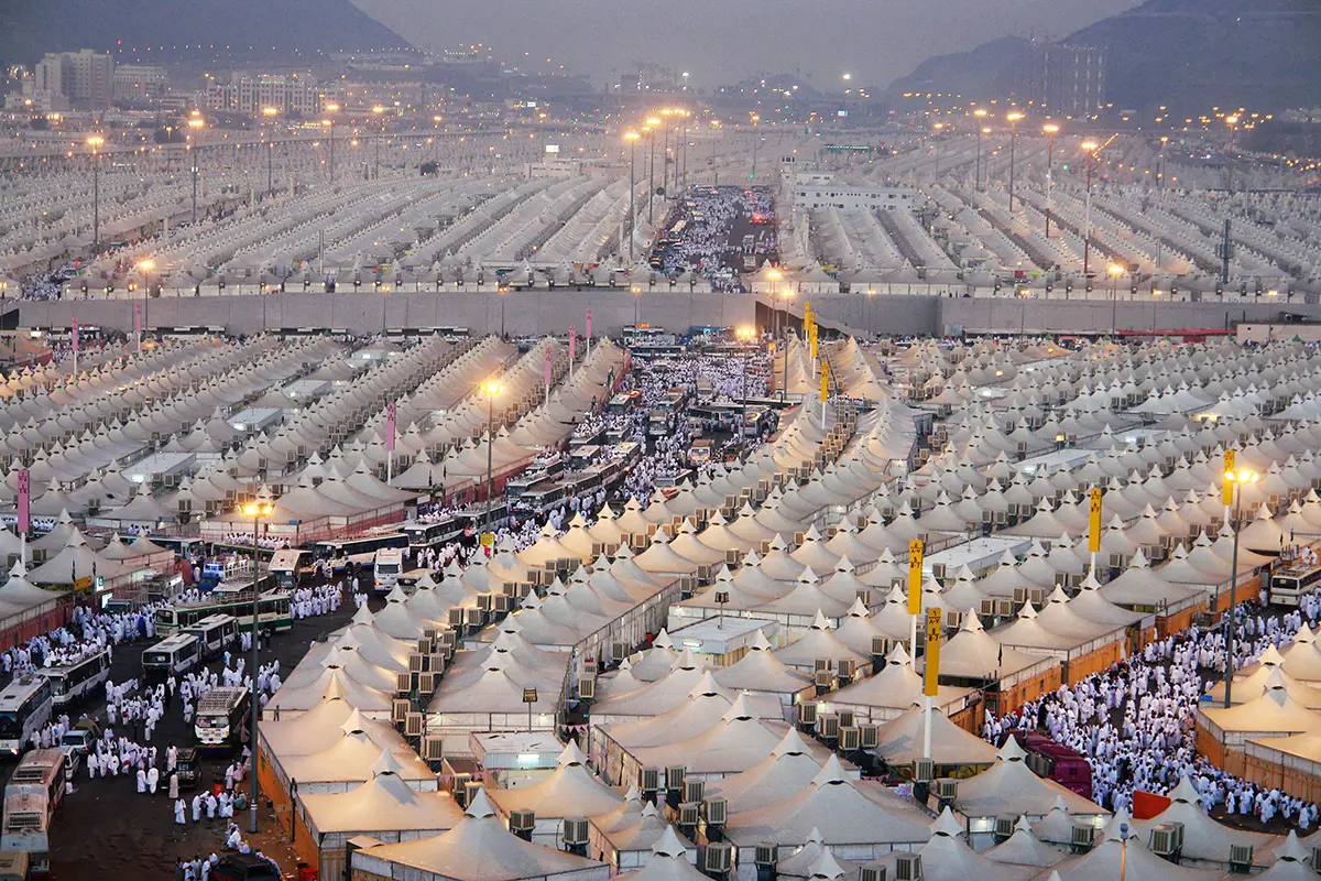 Empty gravel ground between rows of white Hajj tents in the Mina valley, low bare mountains behind, no building standing where the old mosque of al-Kawthar once was.