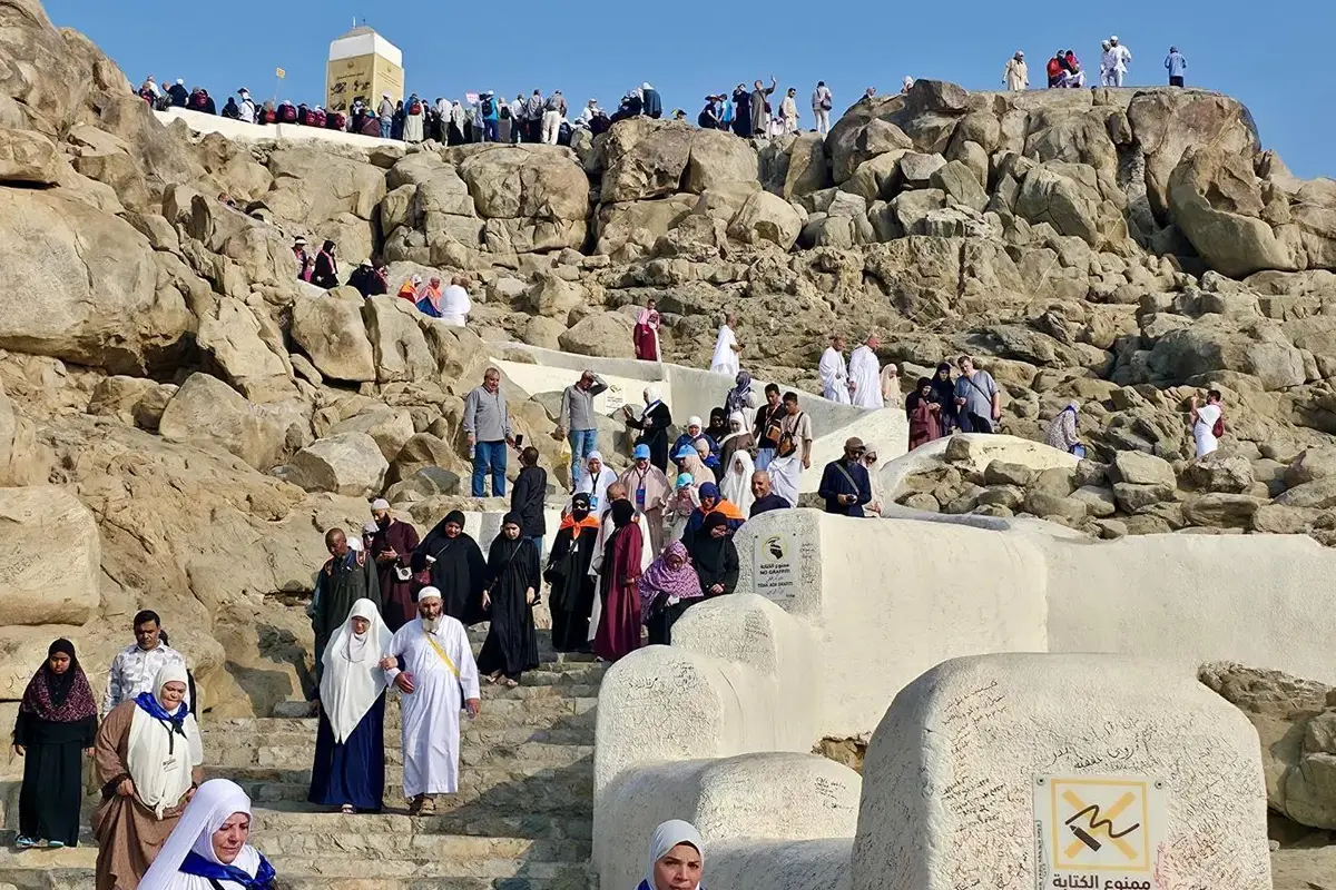 The rocky lower slope of Jabal al-Rahmah on the plain of Arafat, large weathered boulders and open gravel ground where Masjid al-Sakharat once stood, the white pillar on the summit above.