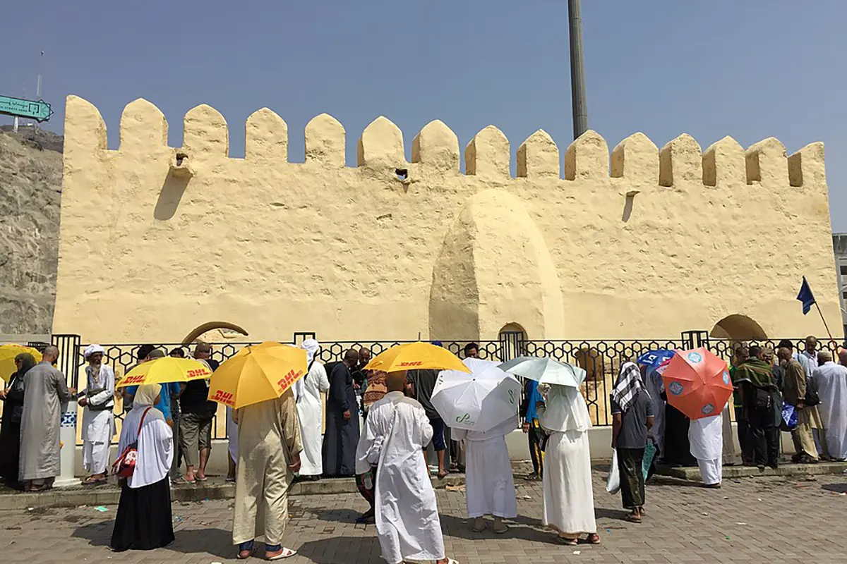 The small roofless stone enclosure of Masjid al-Bay'ah near Jamrat al-Aqabah in Mina, low ancient walls and a plain mihrab niche, modern Hajj infrastructure rising immediately behind it.