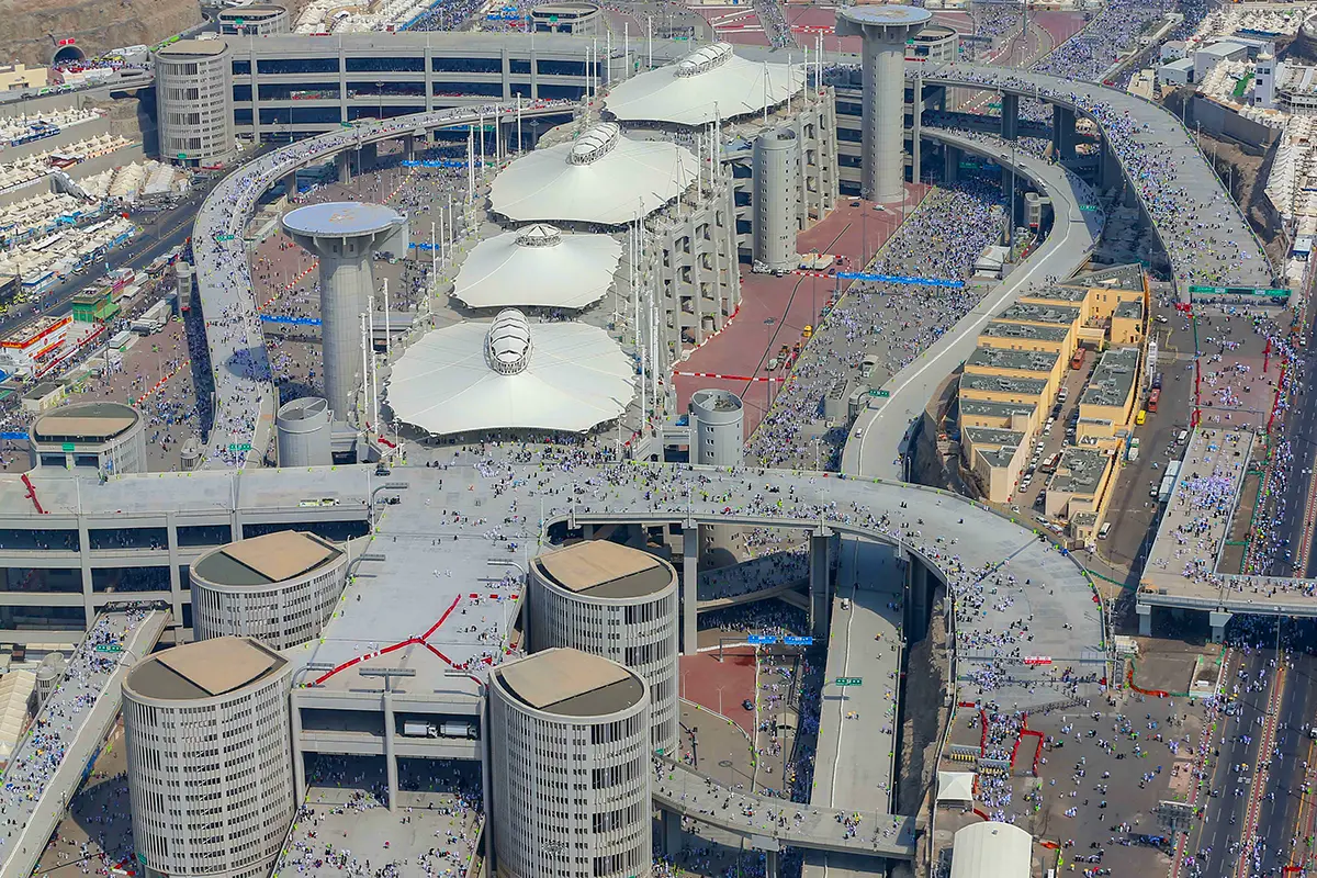 The multi-level Jamarat complex in Mina, its long concrete decks and ramps stacked above one another, pilgrims moving across the upper floor as anonymous blurred shapes towards the oval stone pillar.