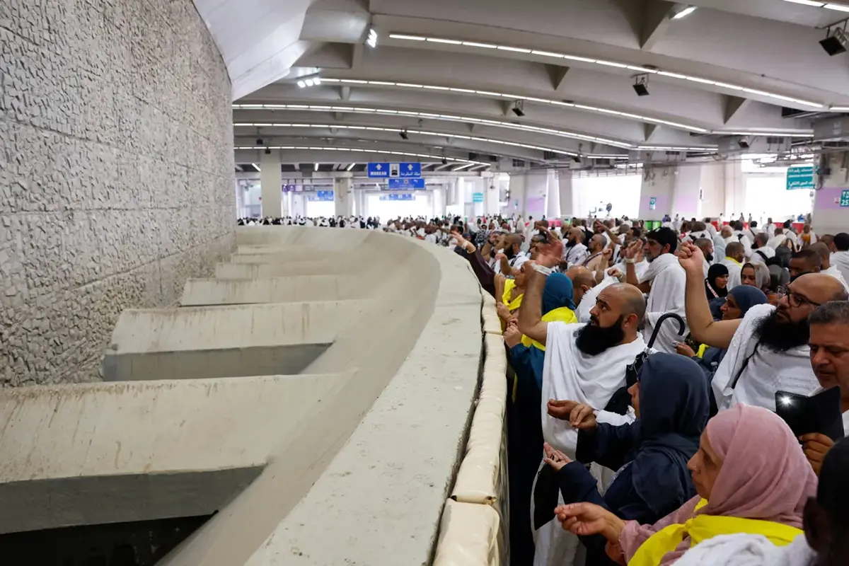 The tall oval stone wall of Jamrat al-Aqabah at the Makkah end of the Jamarat complex in Mina, its wide catch basin below, pilgrims throwing from the upper deck as anonymous blurred figures.