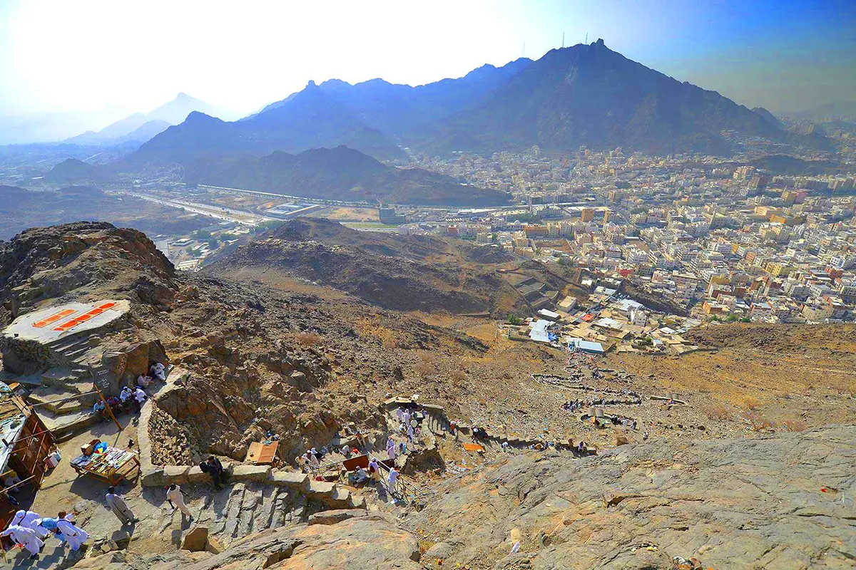 The rocky slope of Jabal al-Sayyidah rising above the walled al-Mu'alla cemetery in the al-Hajun district of Makkah, apartment blocks stacked up the hillside and a ring road curving below.