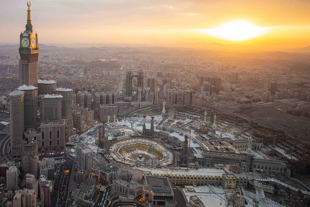 The steep rocky flank of Jabal Abu Qubais rising directly behind the Grand Mosque in Makkah, a large white palace complex on its summit, the mosque colonnades and marble courtyard below.