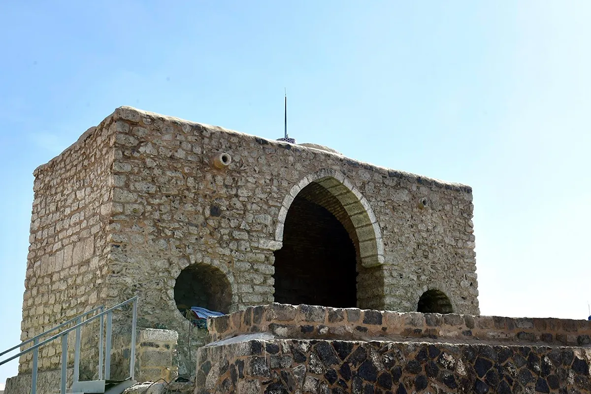 The small pale stone Masjid al-Fath in al-Jumum outside Makkah, a low prayer chamber and short minaret set back from the Madinah highway, dry open ground and brown hills behind it.