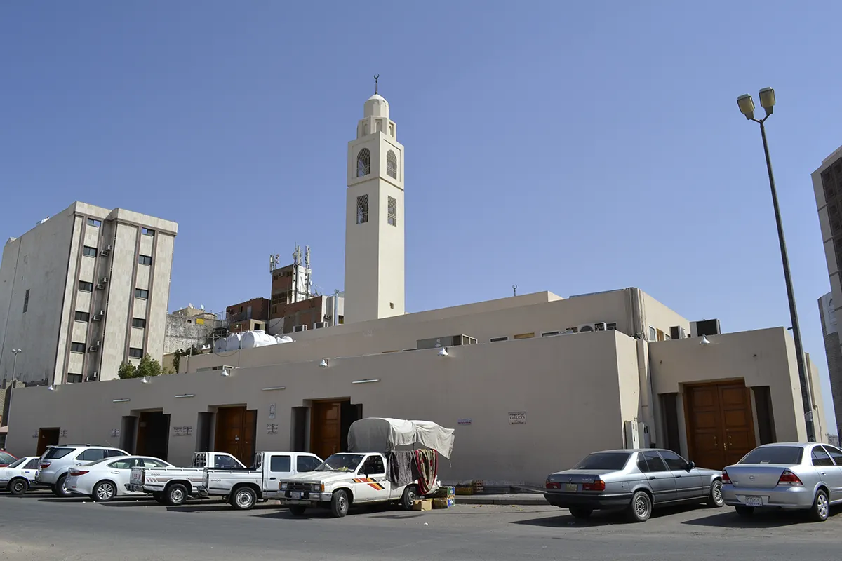 The small marble-clad Masjid al-Ijabah at al-Ma'abada in Makkah, a single dome and slim minaret above pale stone walls, apartment blocks and a busy road close on either side.