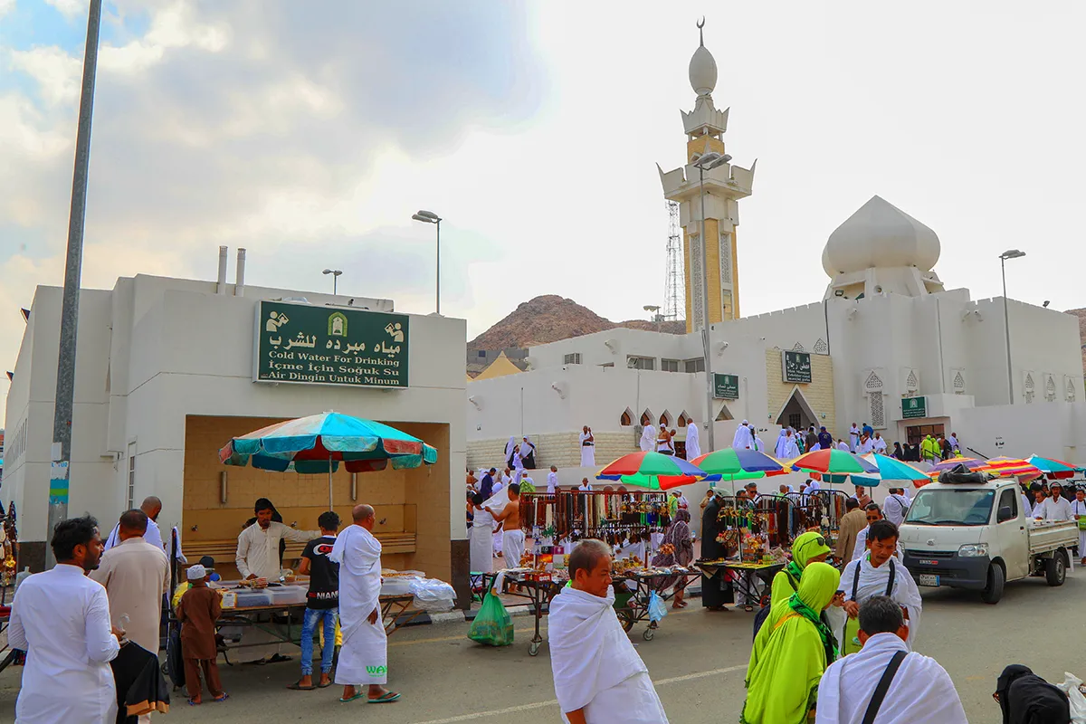 The small white mosque of al-Ji'ranah standing alone in the dry valley north-east of Makkah, a single low minaret against bare brown hills, a few cars parked on the gravel outside.